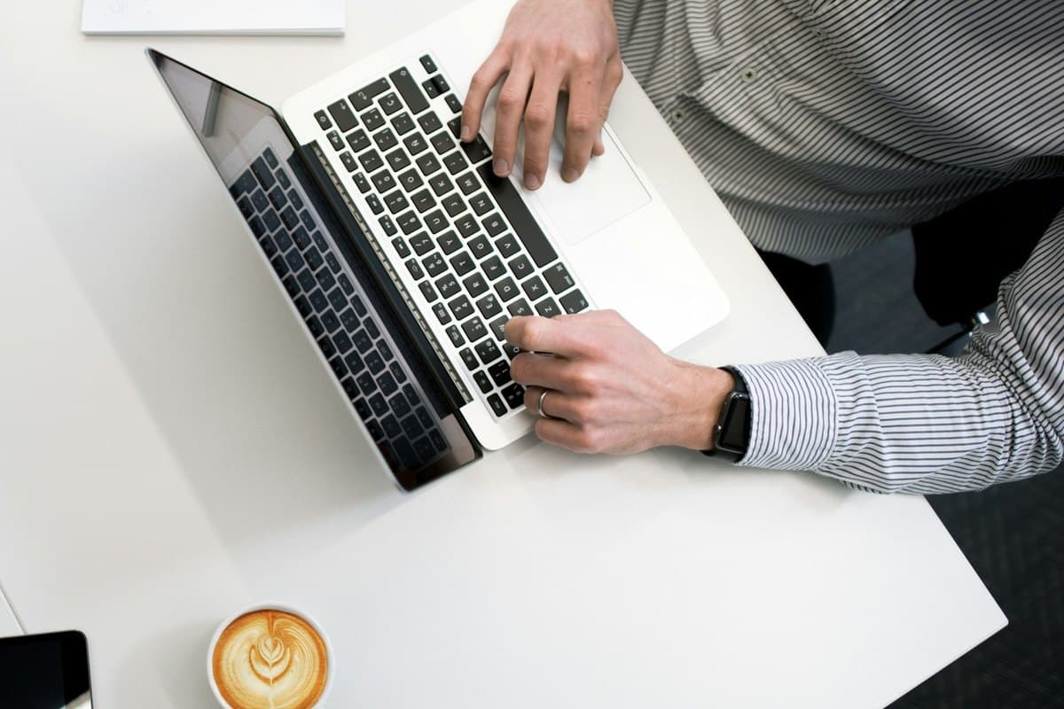 Vue de dessus : une personne travaille sur son ordinateur portable, un café à côté, sur un bureau blanc.