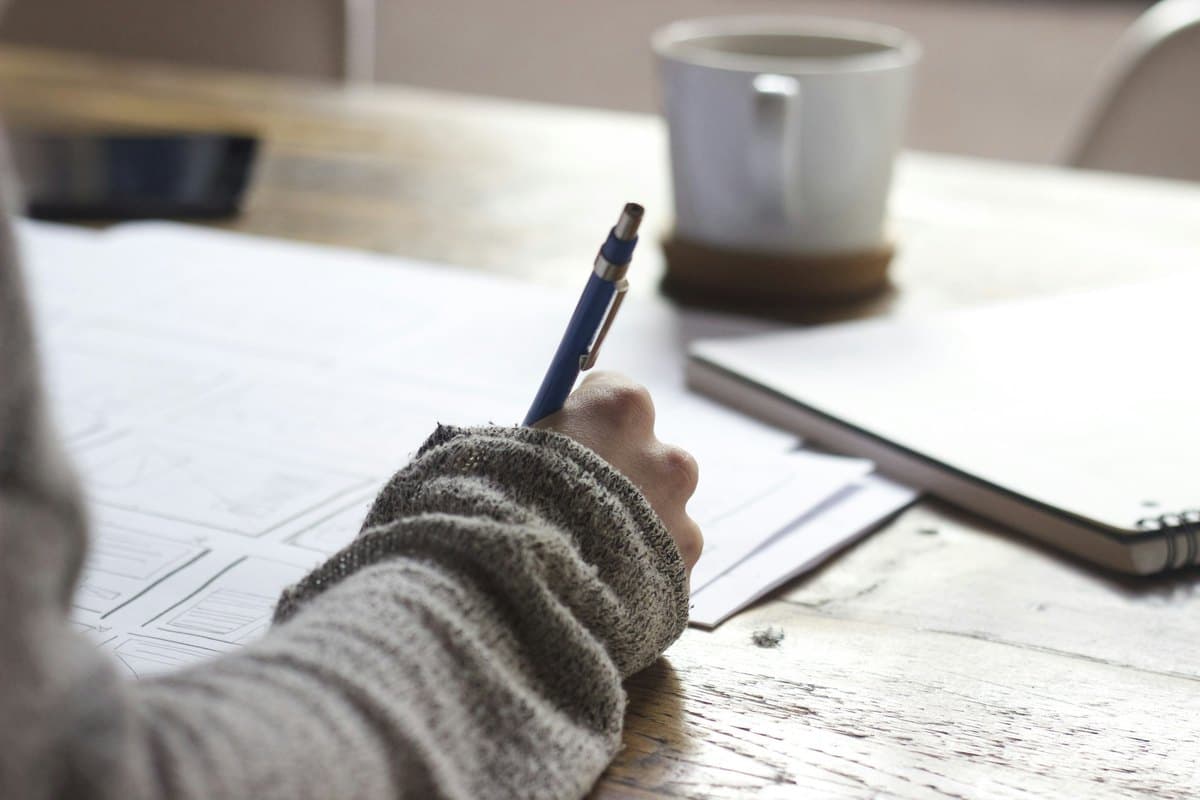 Une main remplit des documents au stylo, une tasse posée sur la table en bois.