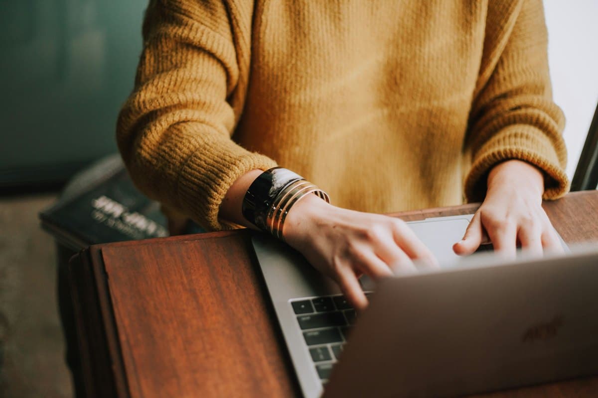 Une personne en pull moutarde tape sur son ordinateur portable, sur un bureau en bois.