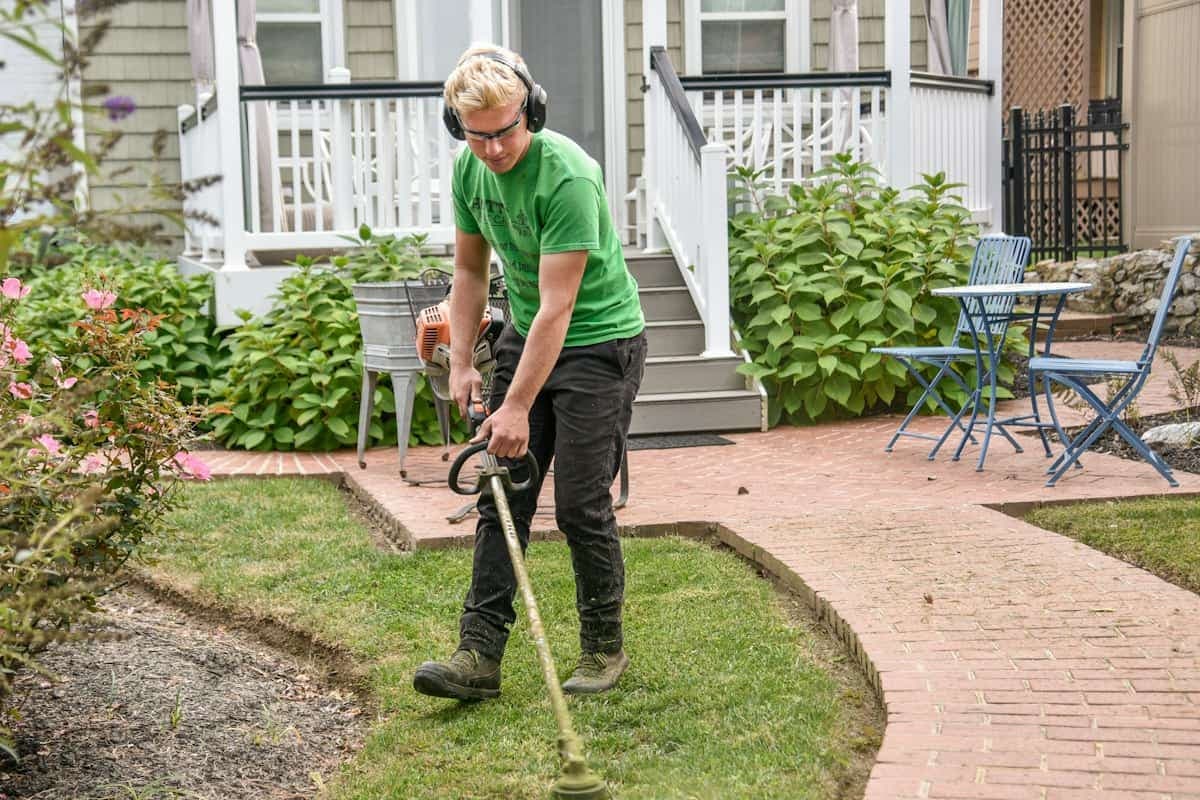 Un jeune homme entretient un jardin avec une débroussailleuse, devant une maison.